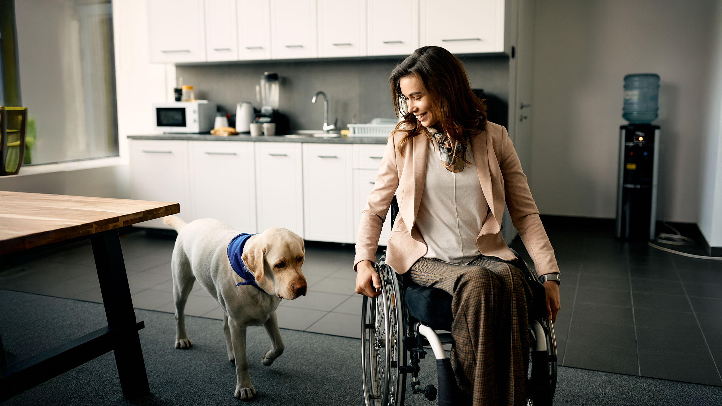 Happy businesswoman in wheelchair and her assistance dog at office kitchen.