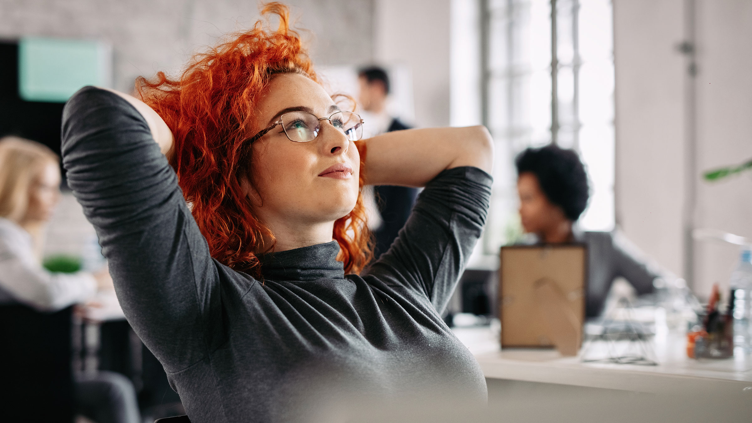 Businesswoman relaxing at desk in the office, tuning out coworkers conversation in the background.