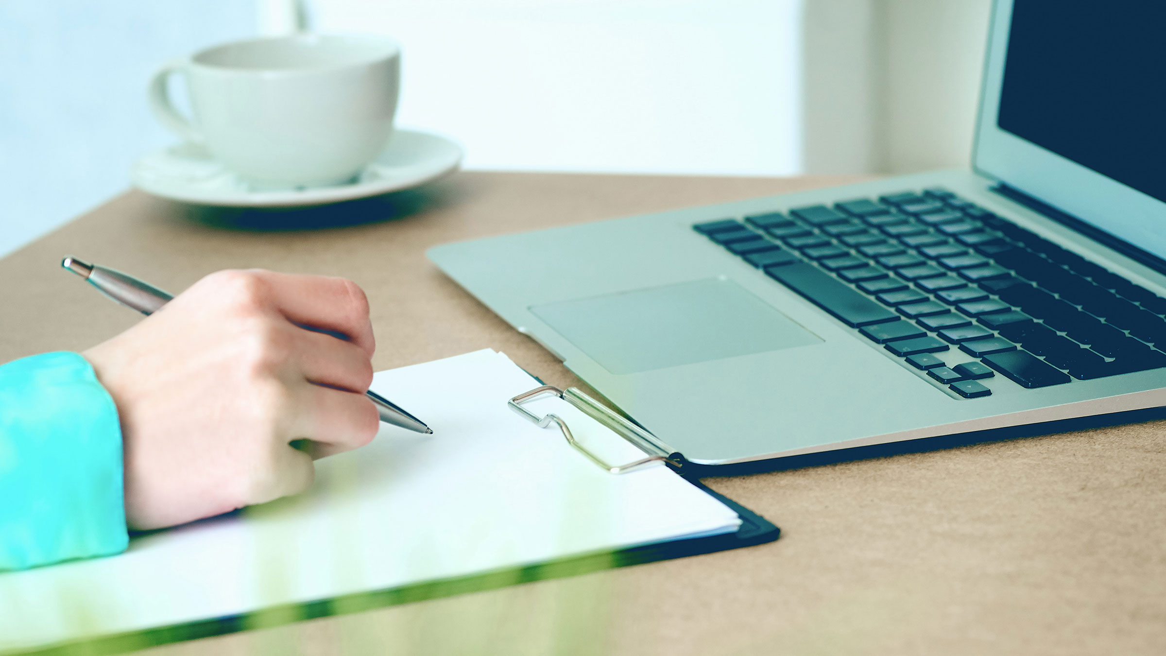 Close-up of female hands writing on clipboard sitting at laptop.