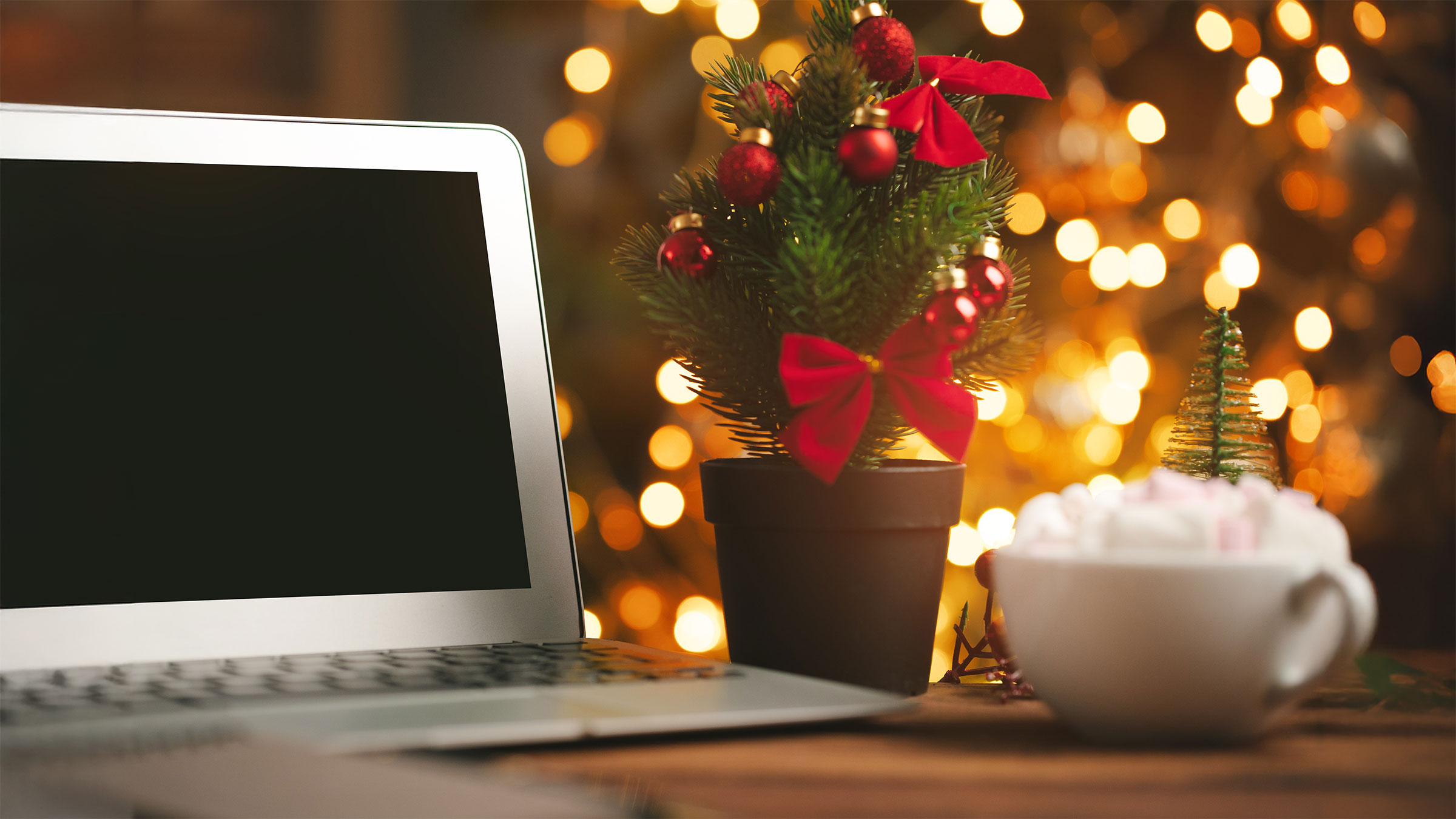 Desk with laptop decorated with small ornamented Christmas tree and blurred lights in background