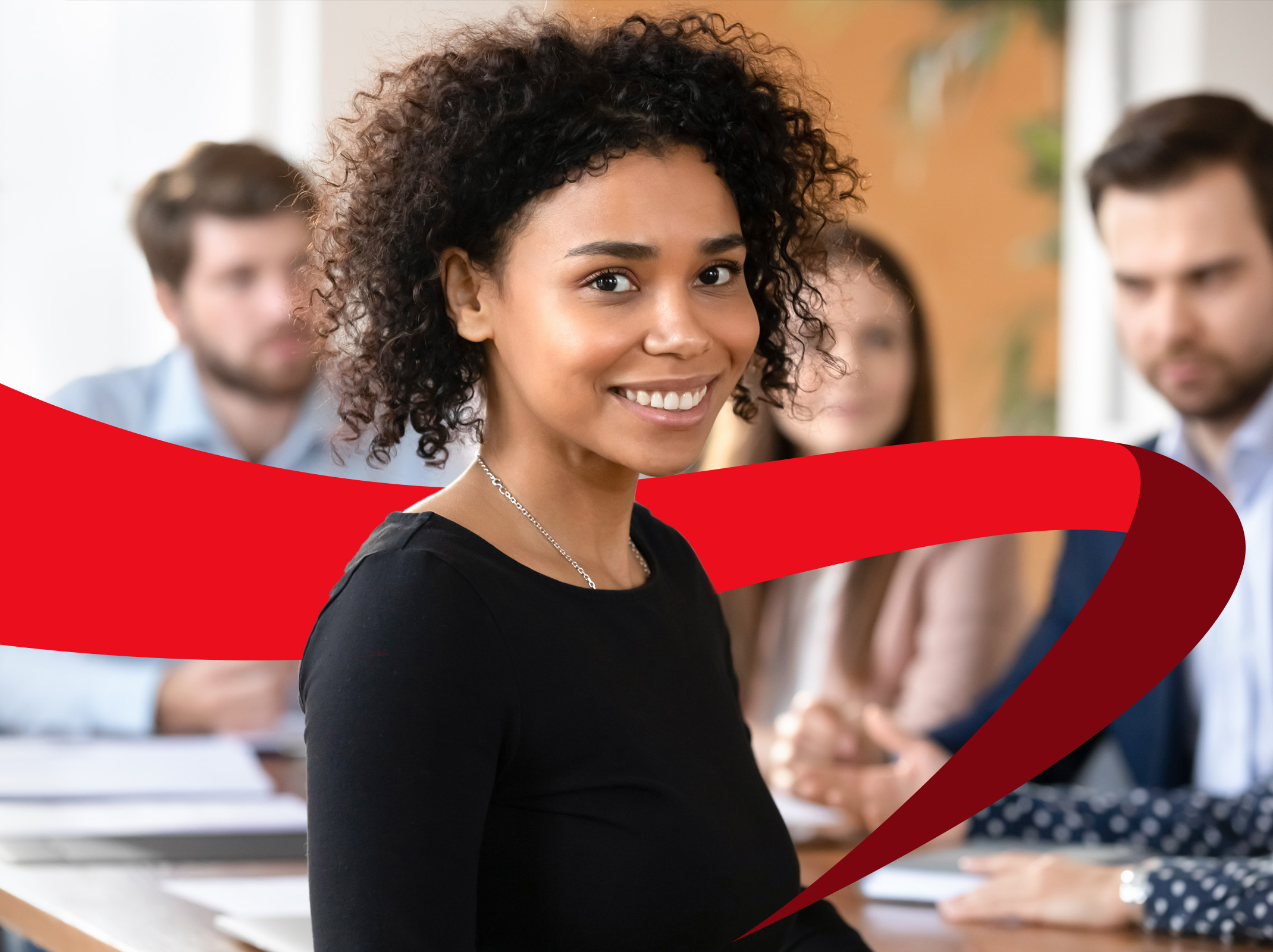 A professional woman smiles directly at the camera while others meet at a table behind her, illustrating workplace success.
