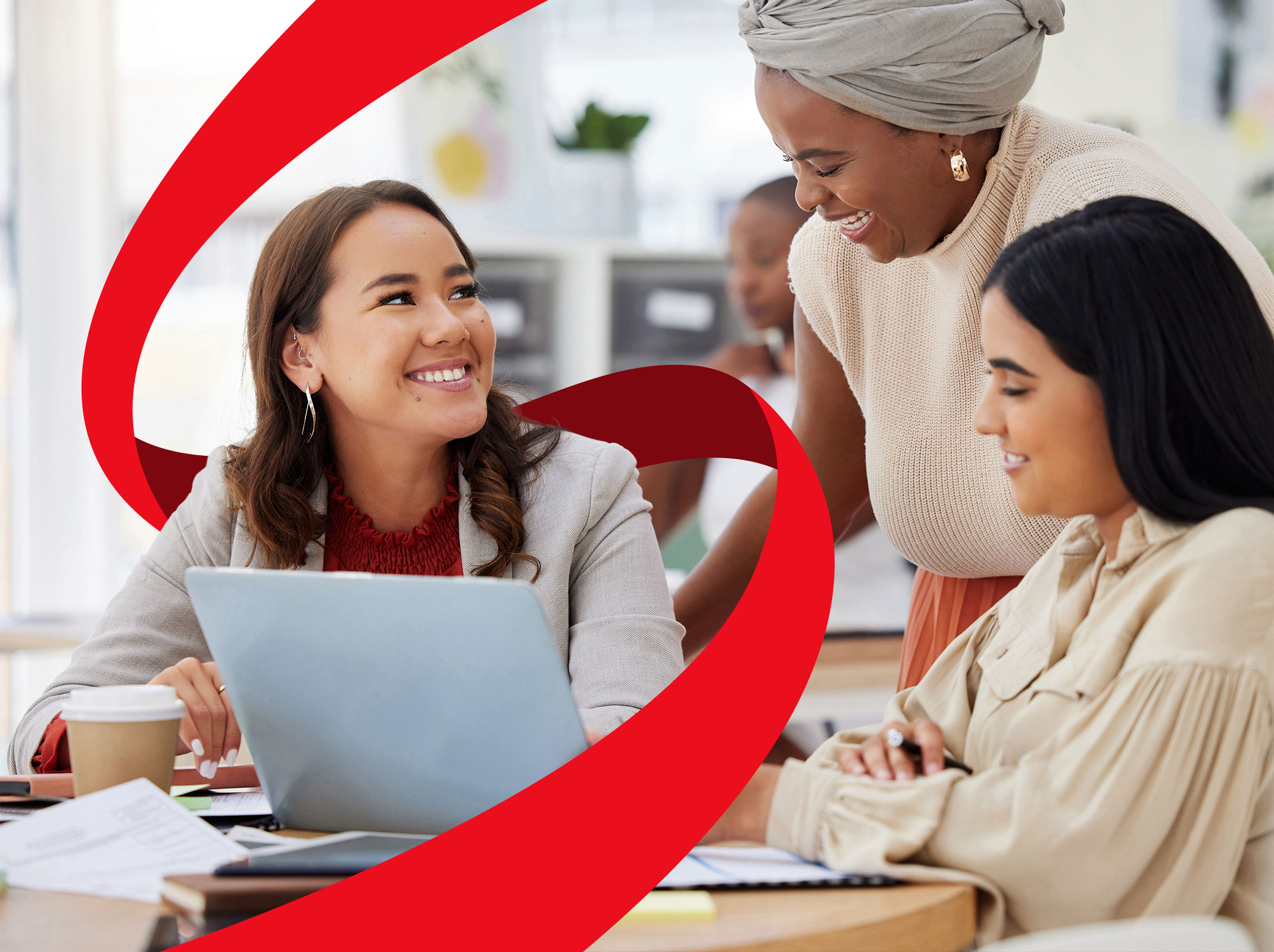 Three professional women in a business setting talking around a computer. They are smiling. One is standing and two are sitting.