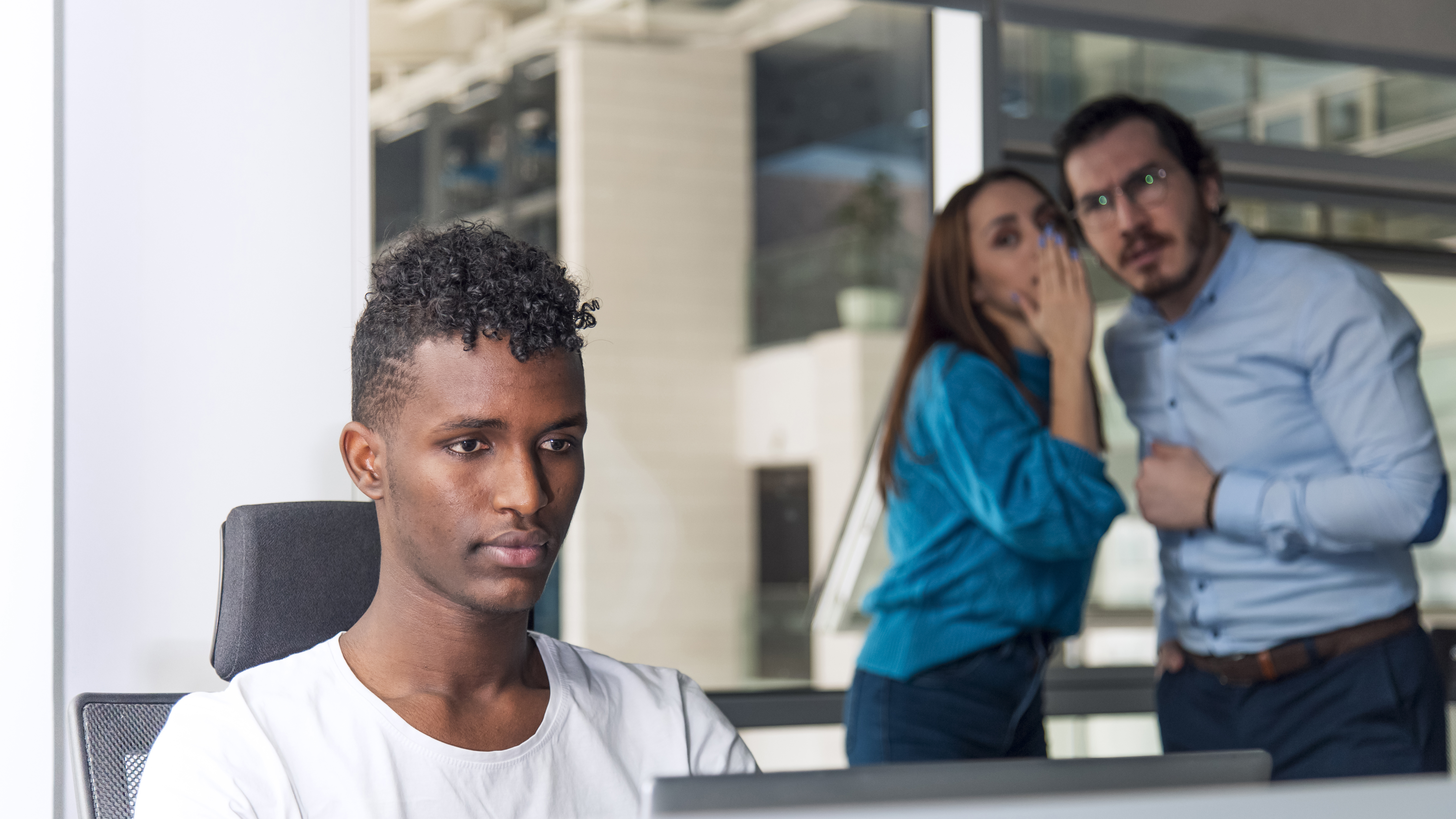 A man in the foreground working while two people in the background whisper