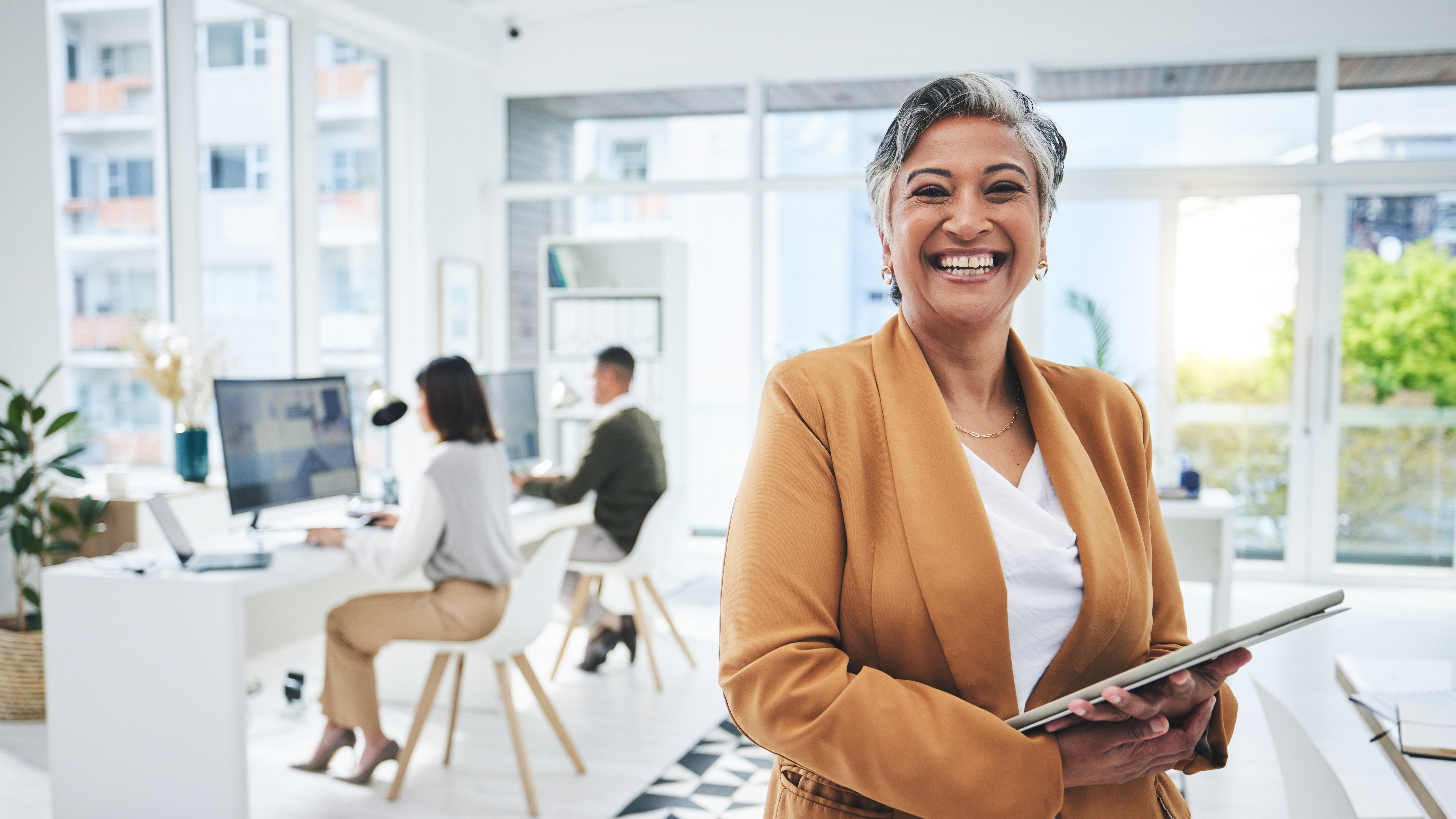 A smiling woman entrepreneur in an office, with two people working at desks behind her