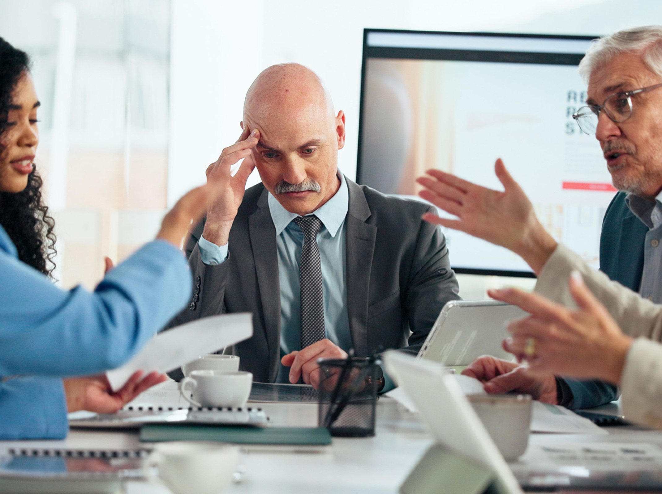 Business people at conference table arguing while man at head of table puts hand at his temple