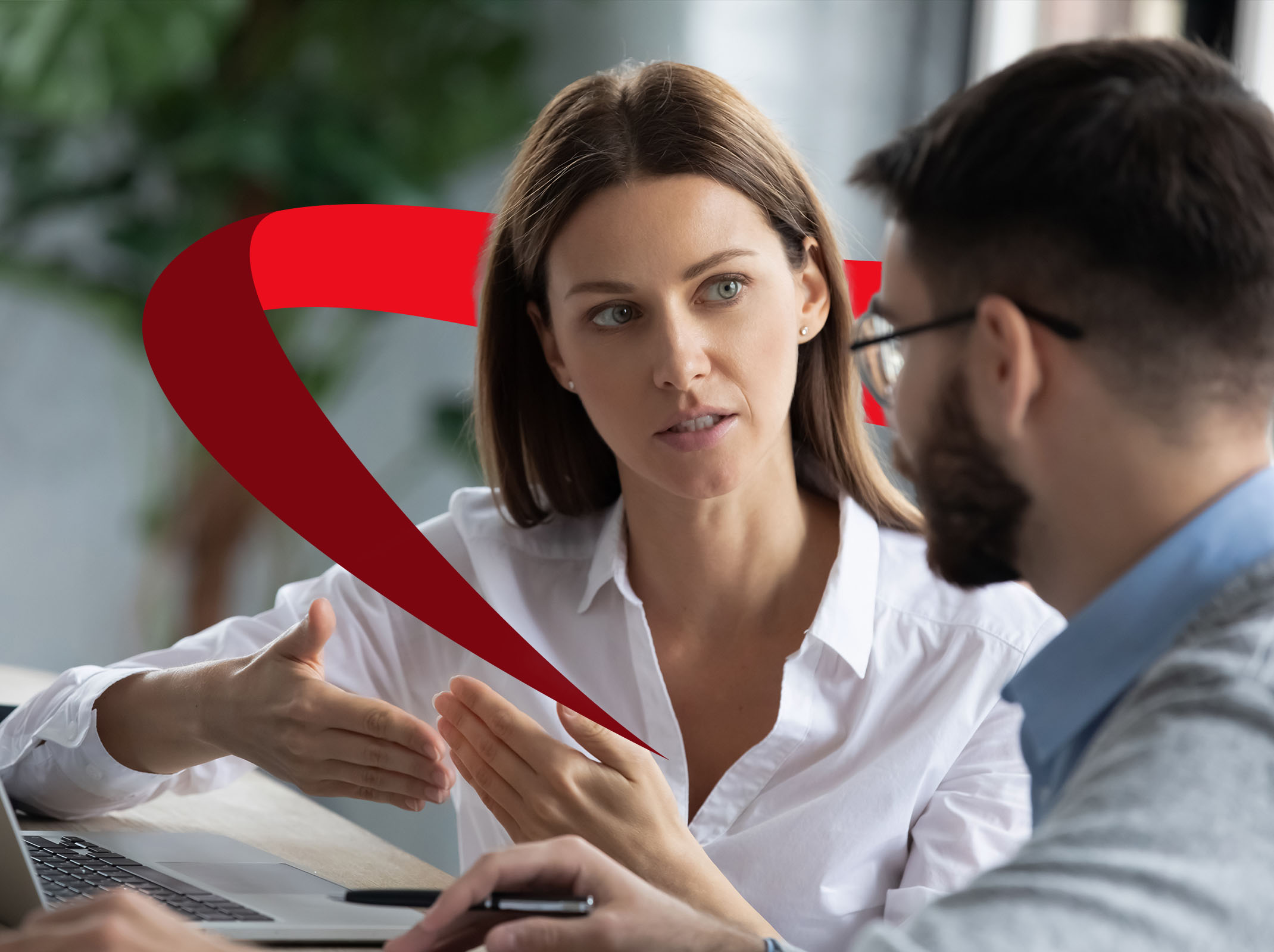 Two colleagues engaged in a face-to-face workplace discussion, using hand gestures while reviewing information at a desk.