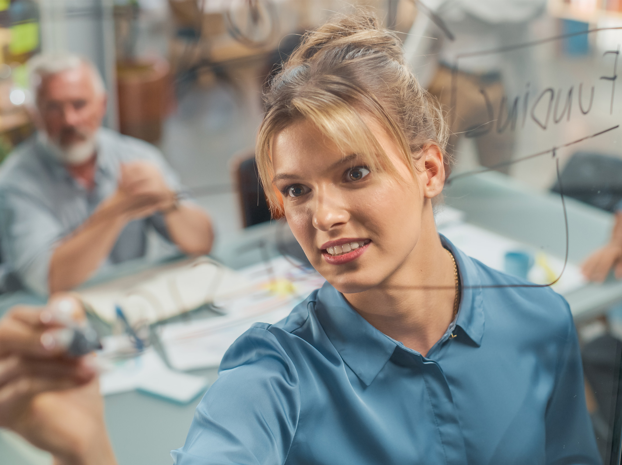 Person in a modern office setting participating in a meeting, leaning forward to write on a glass board while a colleague sits in the background