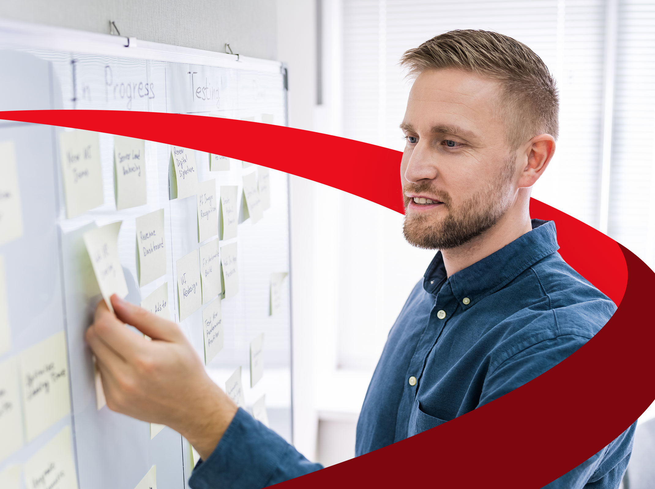 A project manager standing in front of a board of tasks. He is moving sticky notes around on the board in a business setting.