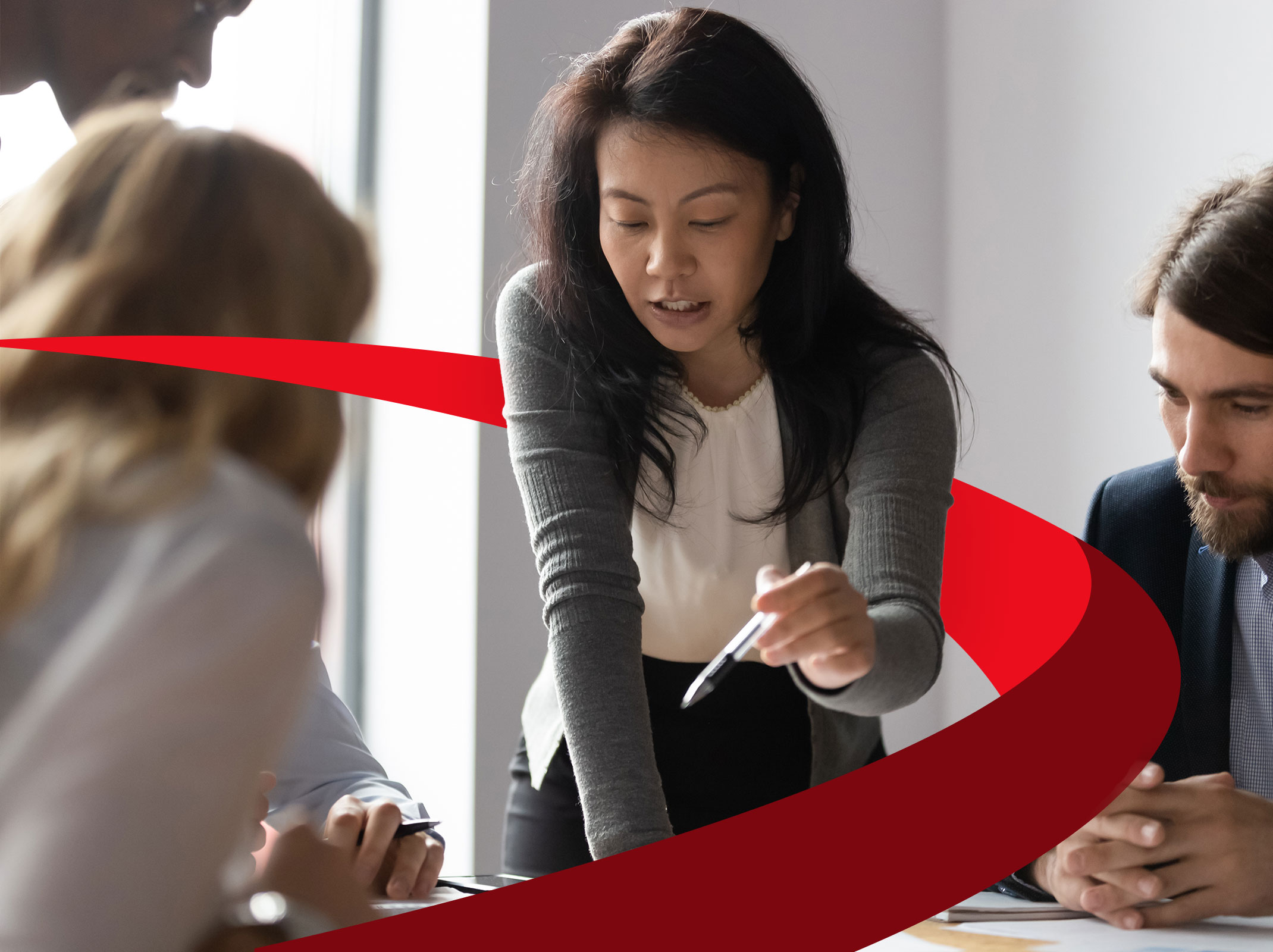 A leader directing a team of two other coworkers around a table in a business setting.  The leader is standing and pointing at something on the table.
