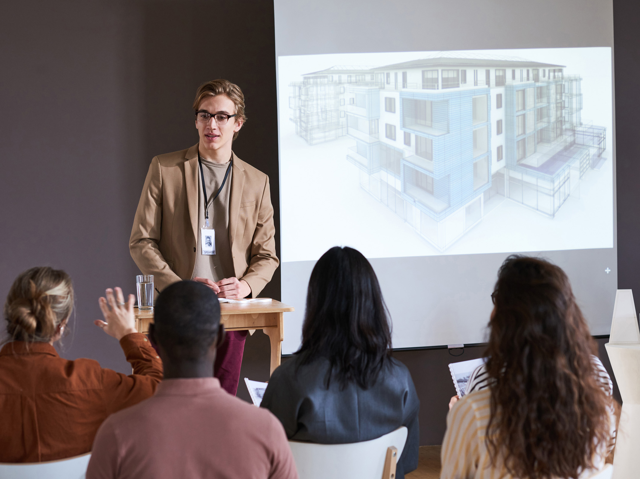 young professional man in suit talking to group with architectural rendering on slide beside him